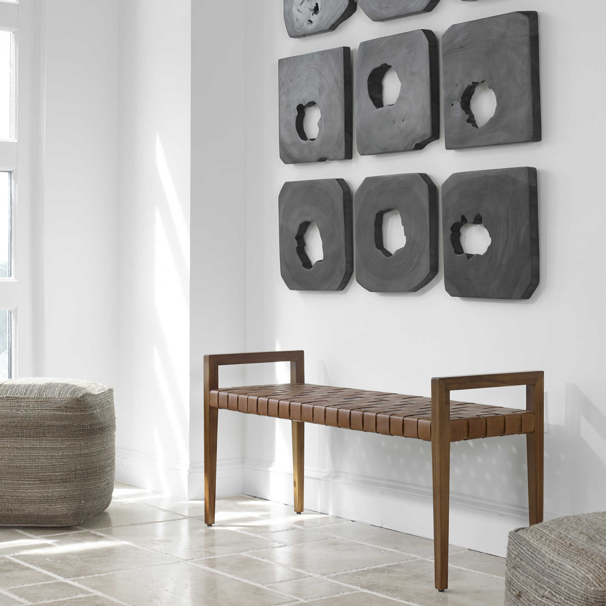 Decorate with leather benches. A plaited leather bench sits in an airy entryway. Soft cube ottomans surround the bench. A large sculptural grid or artwork hangs on the wall above it. 