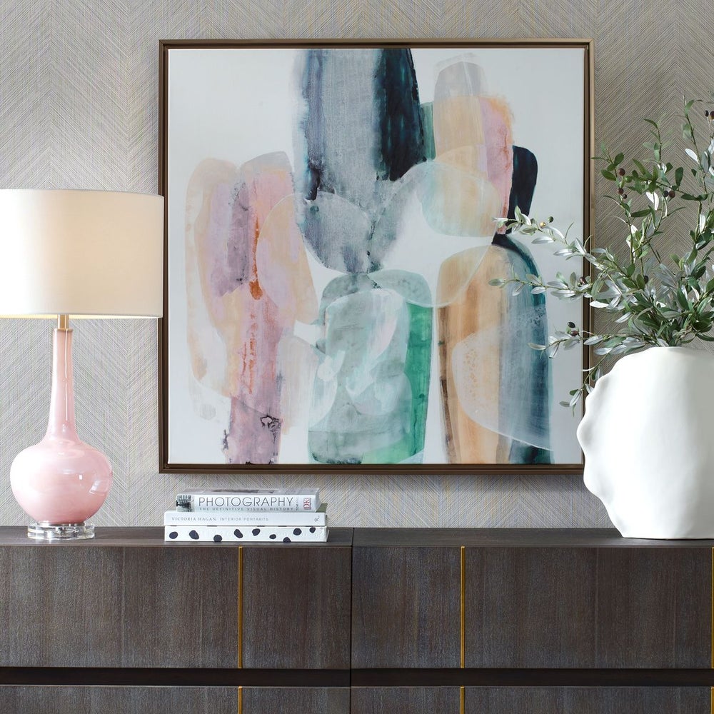 Modern abstract artwork with pink, blue, and green hues above a dark wood dresser, styled with a pink lamp, stacked books, and a white sculptural vase with greenery.