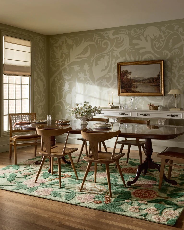 Elegant dining room with pink decor, featuring a floral rug with pink accents, a wooden dining table, vintage chairs, and a neutral wallpapered wall with classic artwork.