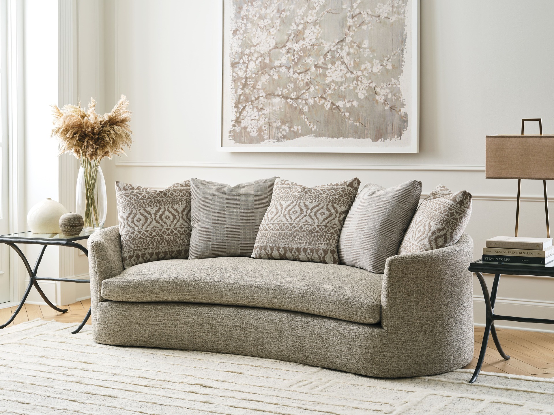 A neutral-toned open concept living room with a curved beige sofa, patterned throw pillows, a soft area rug, and delicate decor elements for a calming and elegant space.