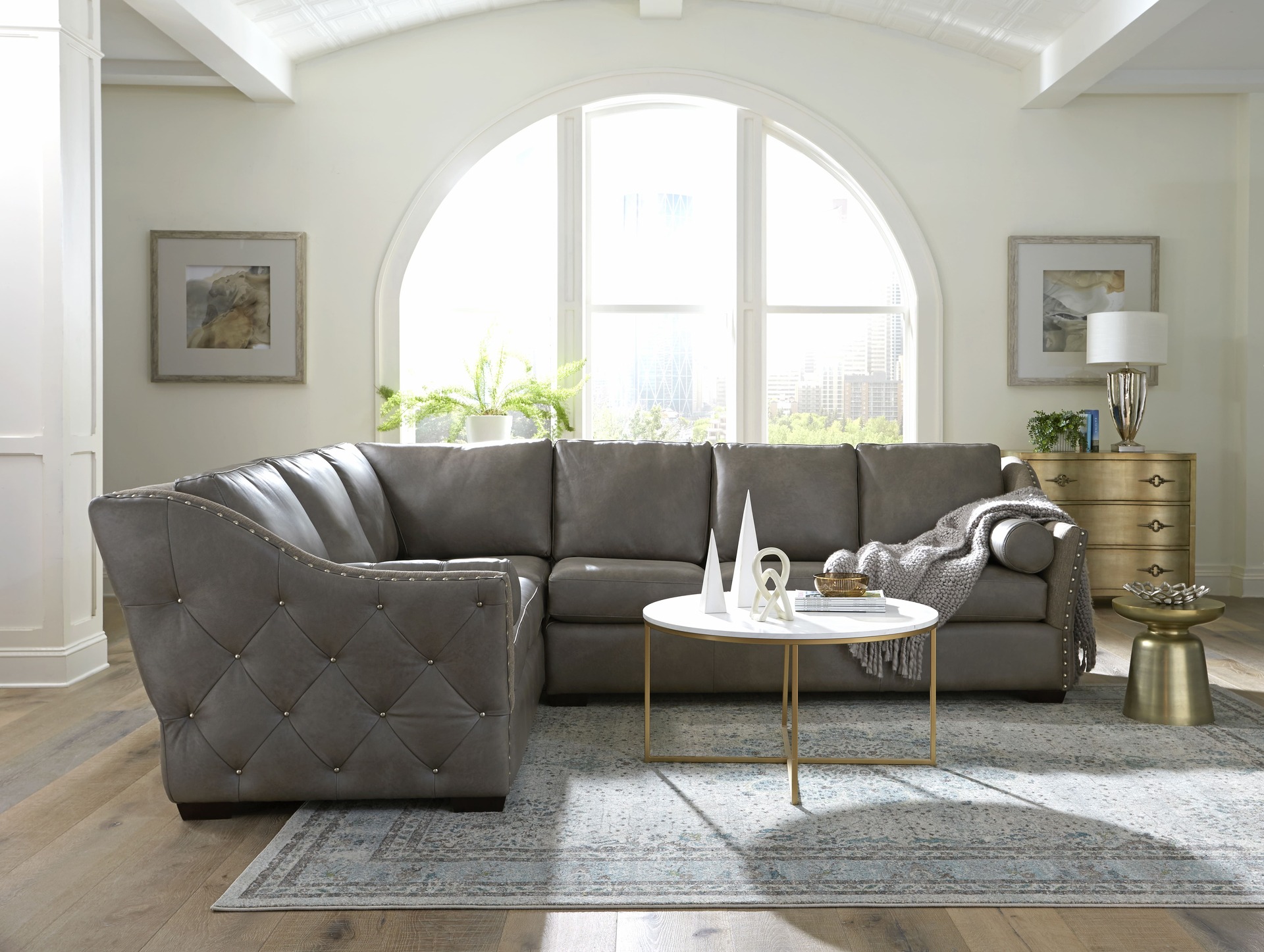 A contemporary open concept living room featuring a gray tufted leather sectional, a sleek white and gold coffee table, and an arched window overlooking the city skyline.