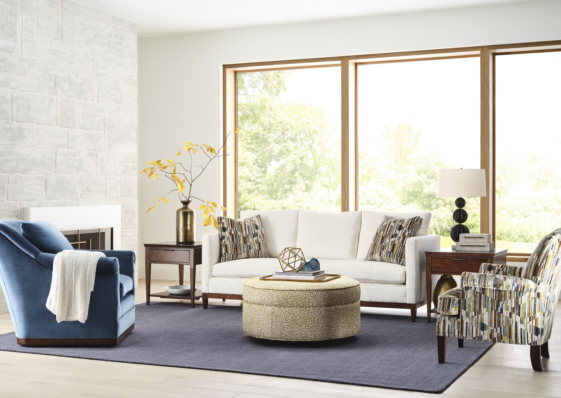 A modern open concept living room with a cream-colored sofa, a navy armchair, a patterned accent chair, and a round ottoman, complemented by large windows and a stone fireplace.