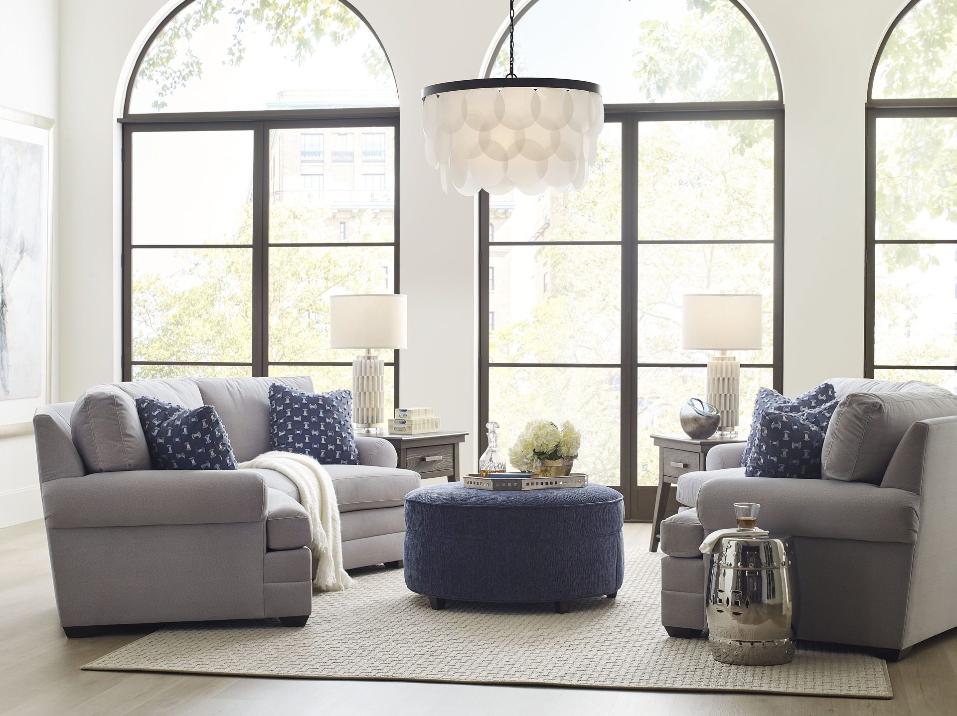 A bright open concept living room with two cozy gray sofas, navy accent pillows, a round blue ottoman, and large arched windows allowing natural light to fill the space.