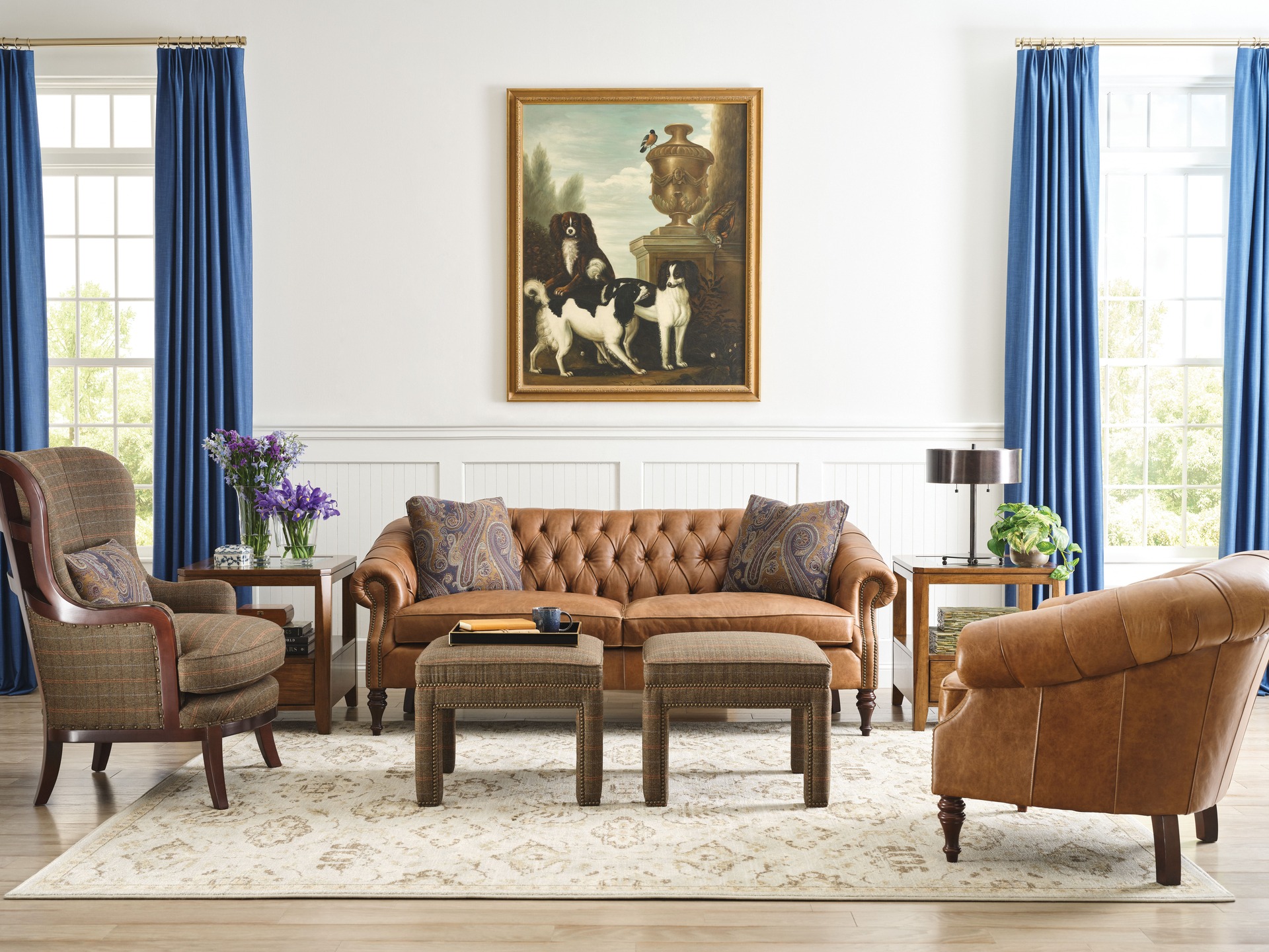 A stylish open concept living room featuring a warm brown leather sofa, a tufted armchair, and rich blue curtains framing large windows, creating a classic and inviting atmosphere.