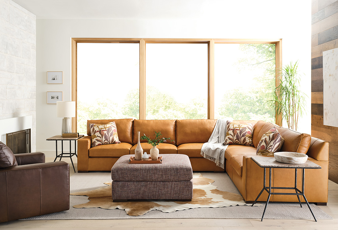 Warm-toned leather sofa sectional in a modern living room with large windows, featuring neutral decor and cozy accents.