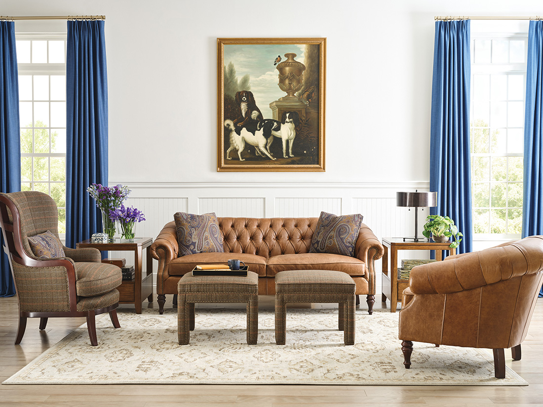 Traditional living room with a tufted leather sofa, matching leather chair, classic wood accents, and blue curtains framing large windows.