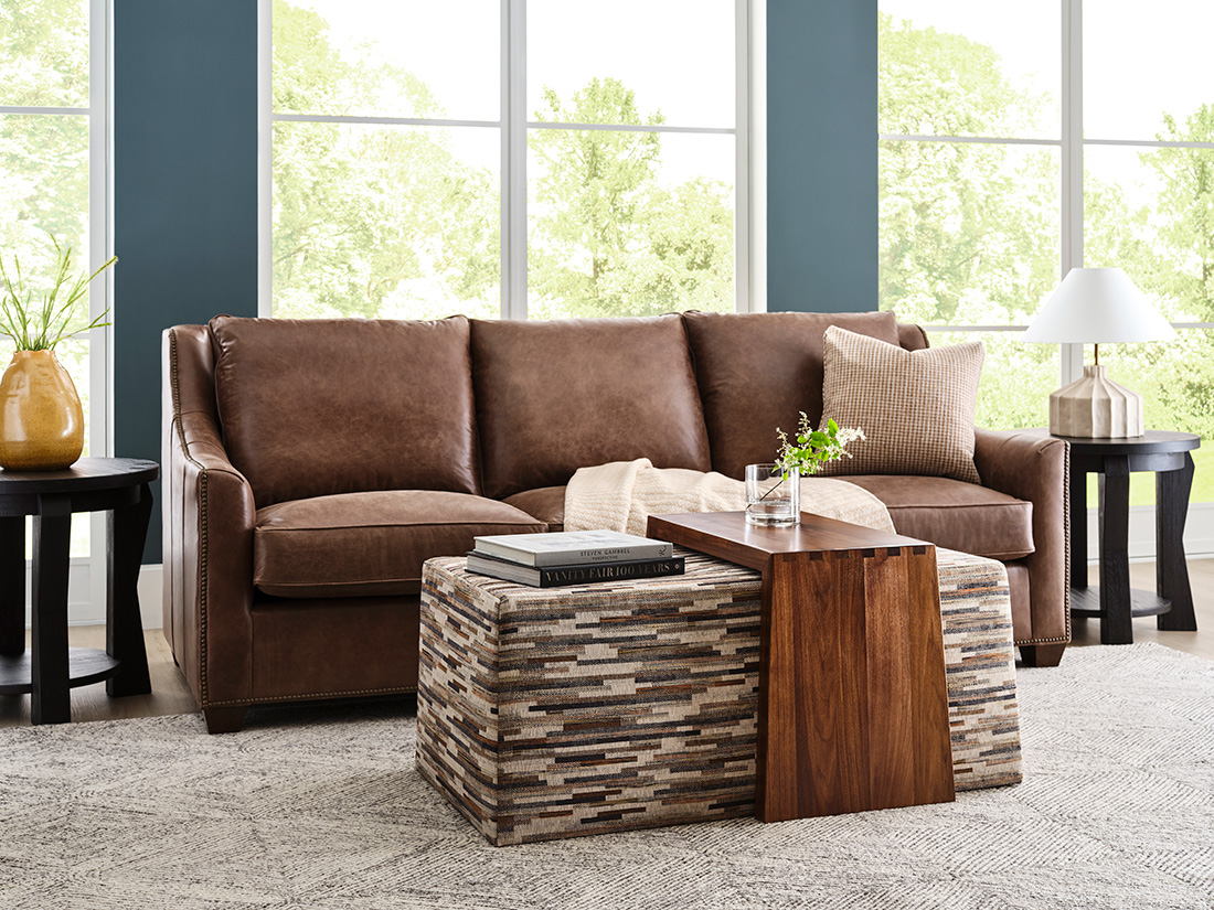 Contemporary living room with a rich brown leather sofa, modern accent tables, and a geometric ottoman, set against large windows and deep teal walls.