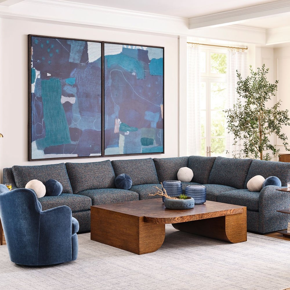 Living room with a large blue sectional sofa, a curved armchair, and a sculptural warm wood coffee table, accented by modern blue artwork and natural light.