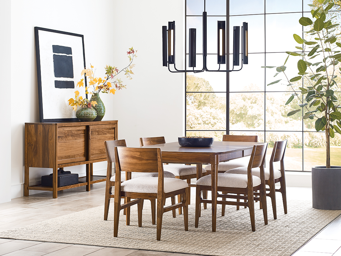 Modern dining room featuring warm wood furniture, including a solid walnut table, matching chairs with neutral cushions, and a sideboard, all set against large grid windows and a statement black chandelier.